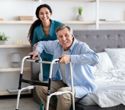 female-nurse-helping-elderly-male-with-walking-frame-stand-up-from-bed-at-home-professional-care.jpg